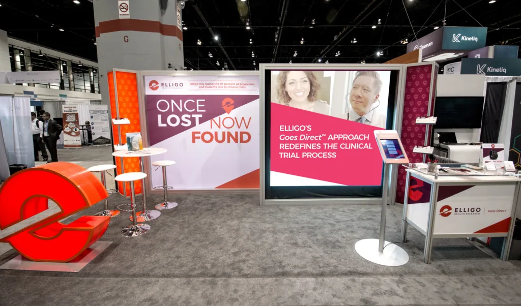 A trade show booth for Elligo Health Research with a bold, modern design. The left side features a large illuminated red “e” logo with a seating area that includes tall white chairs and a high table. The central wall displays the message “Once Lost, Now Found” with images of a smiling woman and a man using telehealth equipment. The right panel reads “Elligo’s Goes Direct™ approach redefines the clinical trial process.” A touchscreen kiosk stands in front, and a counter with brochures and branded materials sits to the right, showing the Elligo logo and “Goes Direct™” tagline.