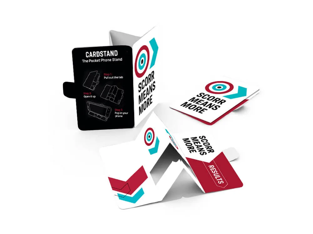 Foldable SCORR-branded pocket-sized phone stands, called “CARDSTAND.” The items are shown flat and assembled, with printed instructions on how to pop them open. The design includes the SCORR bullseye logo and the phrase “SCORR MEANS MORE.”