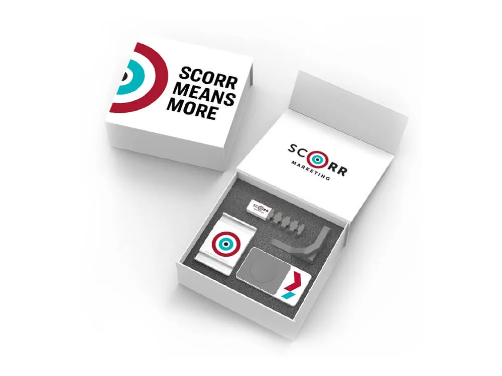 A branded SCORR Marketing desktop essentials kit packaged in a white box labeled "SCORR MEANS MORE." The open box reveals items such as a phone stand, USB drive, microfiber cloth, and a few small accessories, all custom-fit into a foam insert.