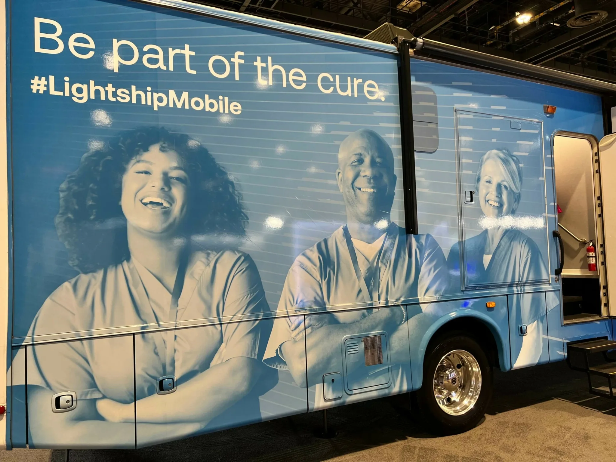 A blue mobile clinical unit featuring large images of three diverse healthcare professionals smiling. The text reads, “Be part of the cure. #LightshipMobile.” The vehicle is parked indoors, with the entrance steps visible on the right side.