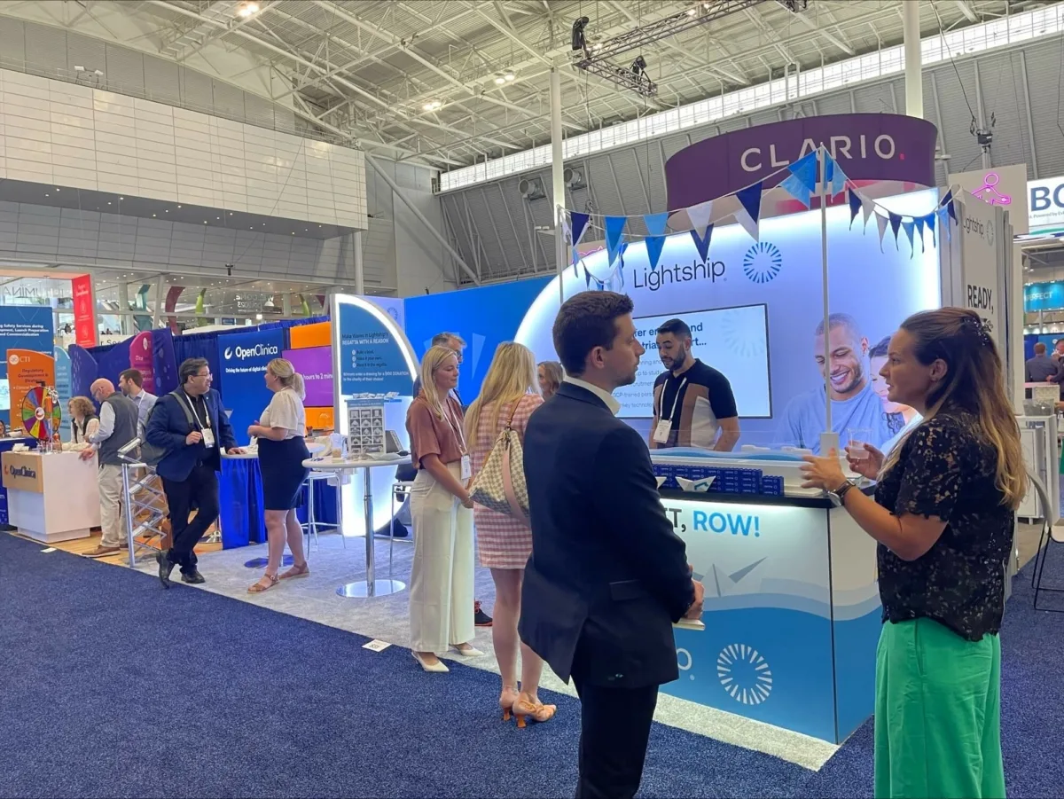 Exhibit hall at a conference with various company booths, including Lightship’s, which is decorated with blue and white bunting. Attendees are seen networking and discussing in front of the booth. Other booths, such as OpenClinica and Clario, are visible in the background.