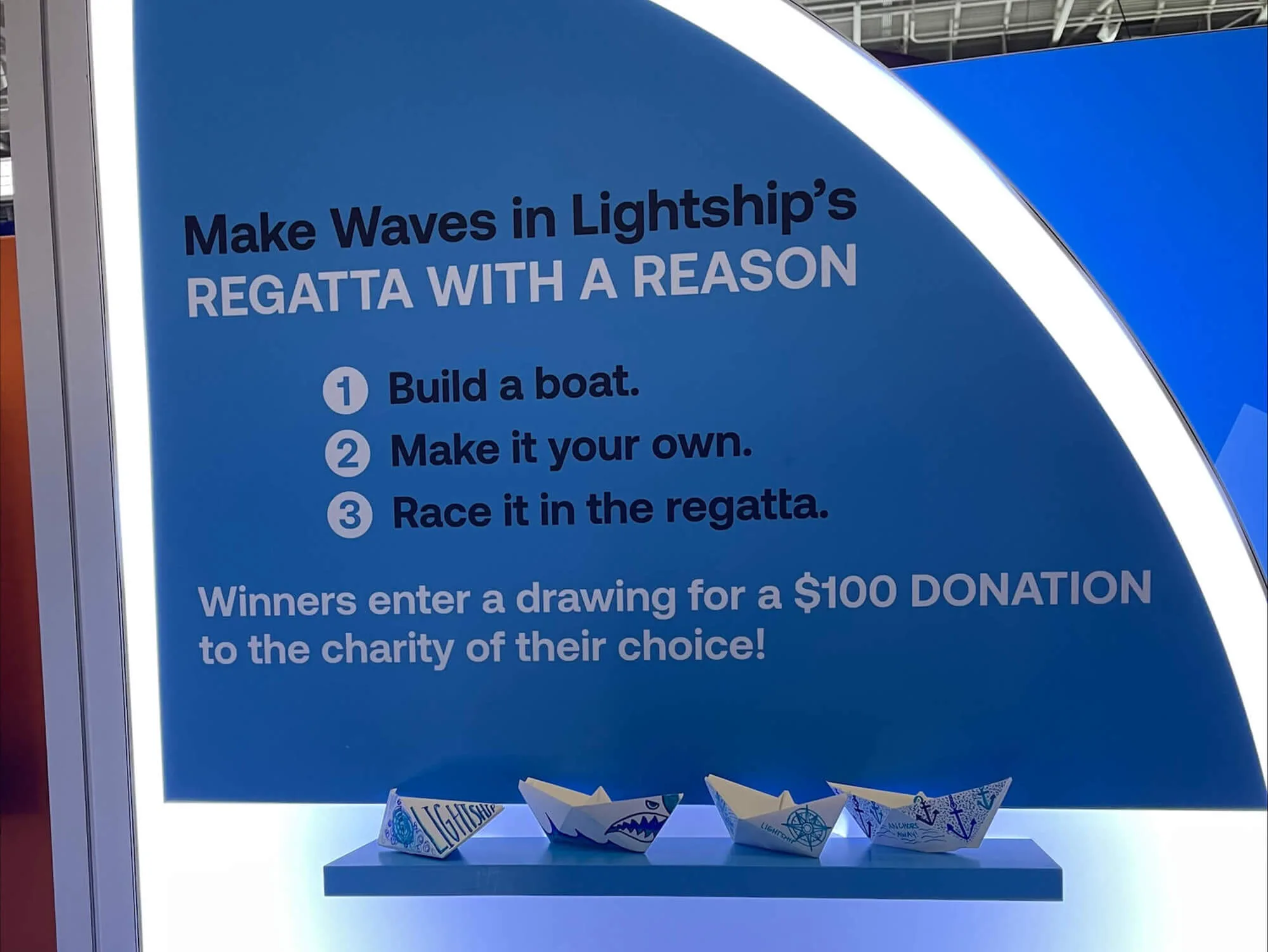 Blue sign at the Lightship booth titled 'Make Waves in Lightship’s Regatta With a Reason.' The instructions read: 1) Build a boat, 2) Make it your own, 3) Race it in the regatta. Winners enter a drawing for a $100 donation to the charity of their choice. Below the sign, several decorated paper boats are displayed on a shelf.