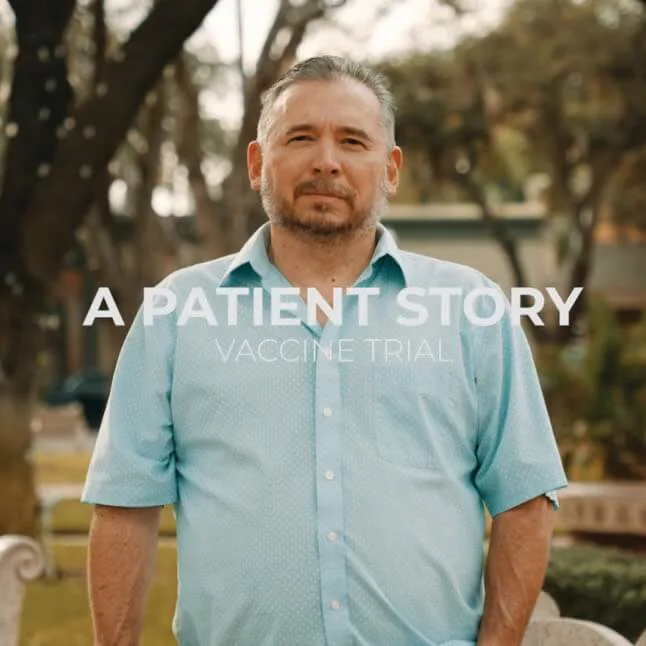 A middle-aged man with short, graying hair and a beard stands outdoors in a park-like setting, wearing a light blue short-sleeved button-up shirt. He has a serious but calm expression. Overlaid on the image is white text that reads 'A PATIENT STORY' with 'VACCINE TRIAL' written underneath in a smaller font.