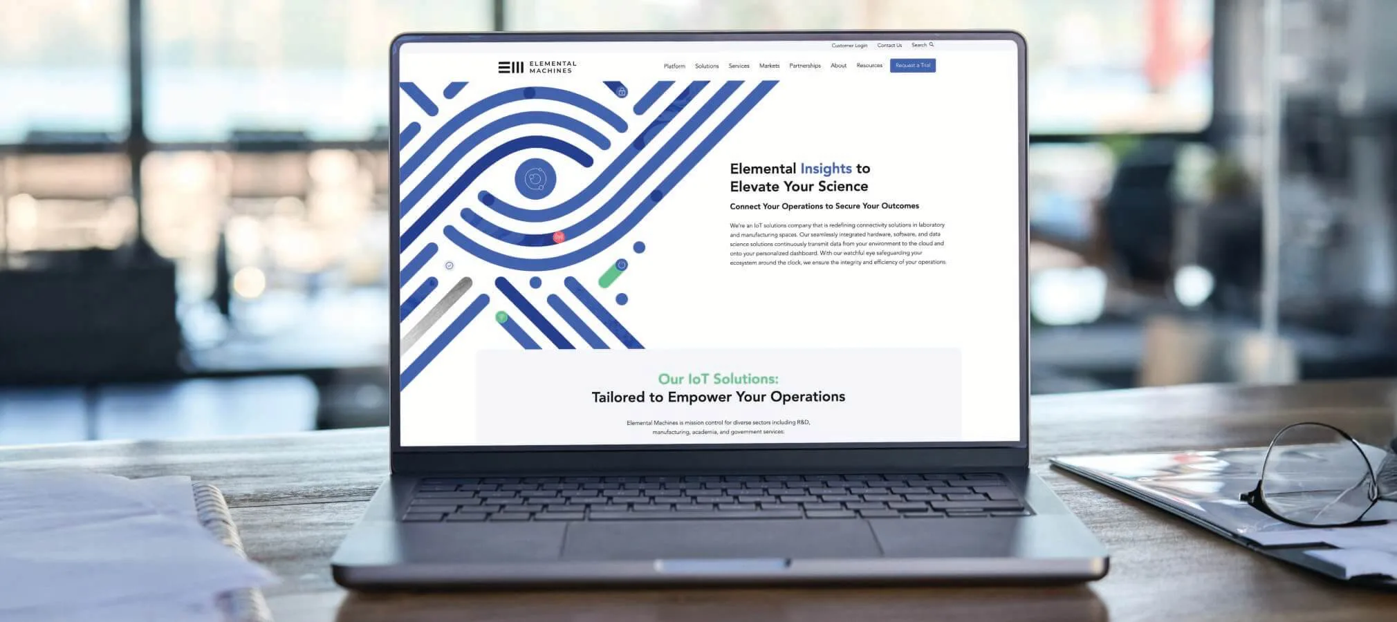 A laptop placed on a wooden table in a modern office setting displays a website for Elemental Machines. The website has a clean design with blue and white graphics resembling circuit lines, and text promoting IoT solutions for laboratory and manufacturing operations. A pair of eyeglasses and some papers are beside the laptop, with a blurred background showing a bright office environment.