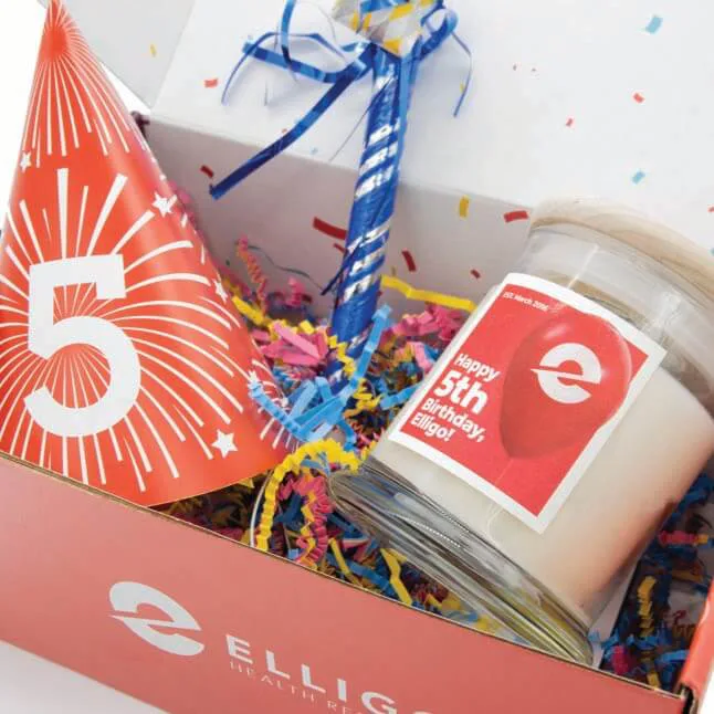 A celebratory gift box for Elligo Health Research's 5th birthday, filled with colorful shredded paper, a red party hat with the number '5,' a blue noisemaker, and a scented candle with a label that reads 'Happy 5th Birthday, Elligo!' The box is branded with the Elligo Health Research logo.