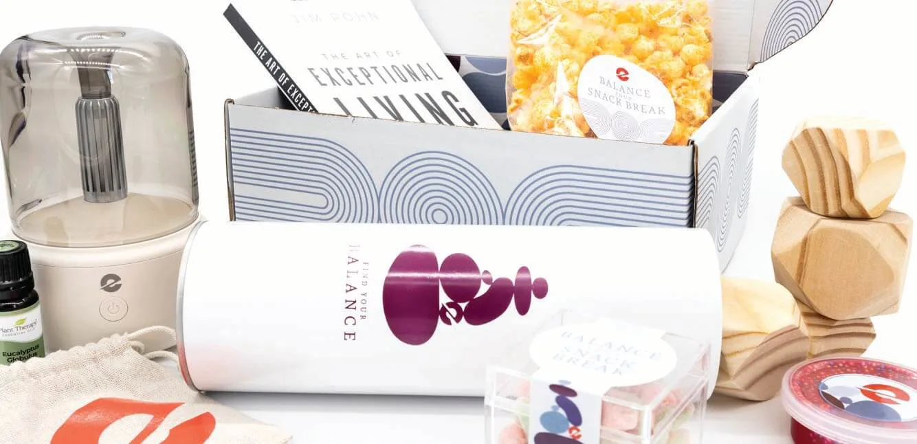 A wellness-themed gift box containing various self-care and relaxation items. The box includes a book titled 'The Art of Exceptional Living,' a small humidifier, a bottle of eucalyptus essential oil, a bag of popcorn labeled 'Balance Snack Break,' a white tumbler with 'Find Your Balance' branding, a drawstring pouch with a red logo, wooden stacking stones, a small container of red putty, and a clear box of colorful gummy candies. The items are arranged in a visually appealing manner with a mix of textures and colors.