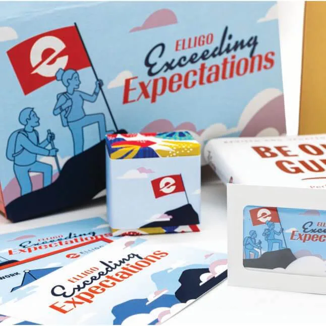 A themed gift set with 'Elligo Exceeding Expectations' branding. The set includes a decorative box featuring an illustration of two hikers planting a red flag with the Elligo logo on a mountain. Other items include a small cube with colorful patterns and the same flag design, a white package labeled 'Be Our Guide,' and various printed materials that reinforce the 'Exceeding Expectations' message. The items are arranged neatly with a cohesive blue, red, and white color scheme.