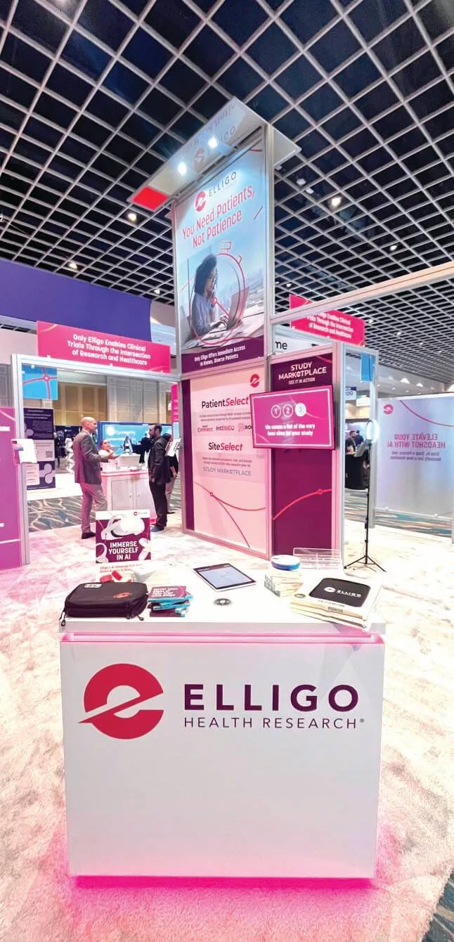 Elligo Health Research trade show booth at a conference, featuring a modern setup with branded signage and promotional materials. A large overhead banner reads 'You Need Patients, Not Patience,' with an image of a researcher using a magnifying glass. The booth includes informational displays about 'PatientSelect' and 'SiteSelect' services, a front desk with the Elligo logo, and various branded giveaways such as brochures, notebooks, and tech accessories. Attendees are seen engaging in conversations in the background.