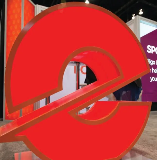 A large, three-dimensional red Elligo Health Research logo on display at a trade show. The logo, shaped like a stylized lowercase 'e' with a diagonal cut, is positioned prominently in the foreground. In the background, trade show booths, banners, and attendees walking around the event space are visible.
