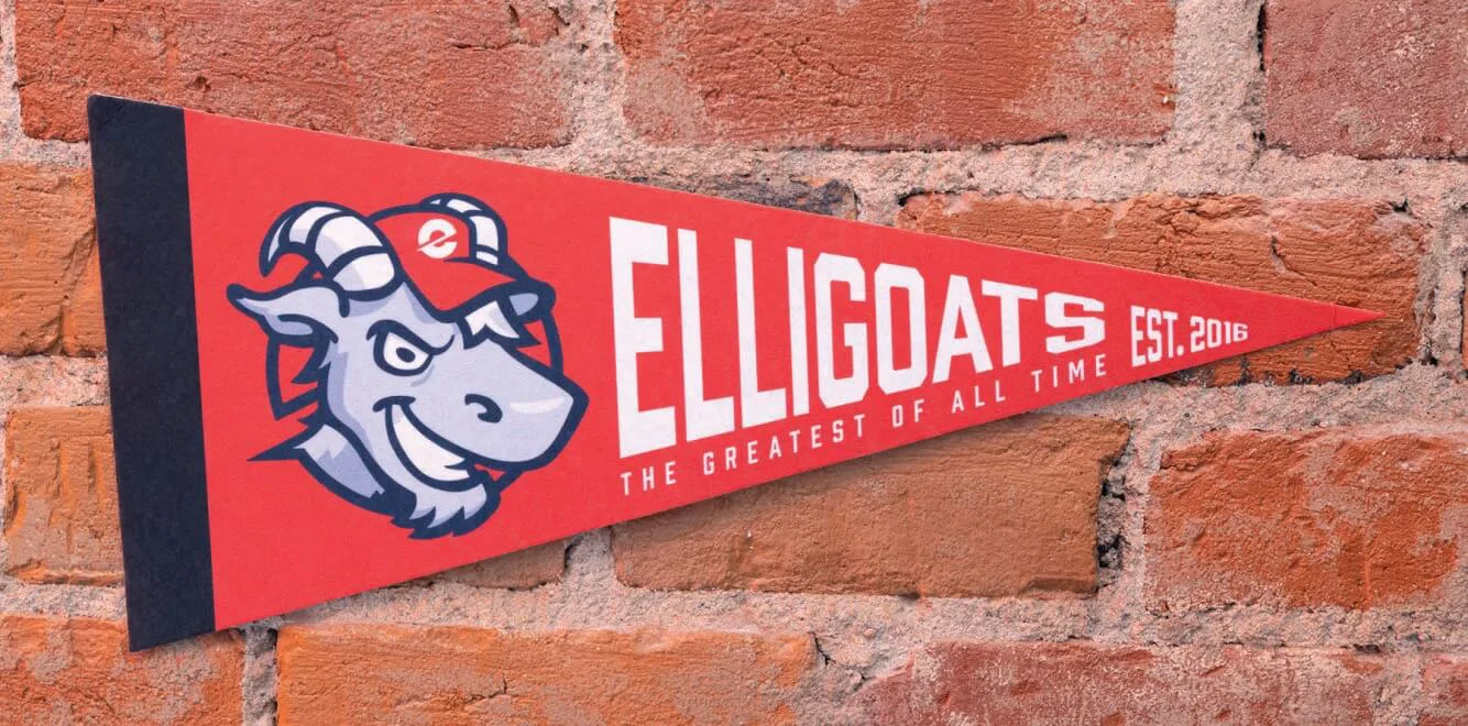 A red and navy blue pennant with the word 'ELLIGOATS' in bold white letters, accompanied by the phrase 'The Greatest of All Time' and 'EST. 2016.' The design features a stylized goat mascot wearing a red cap with the Elligo logo. The pennant is mounted on a rustic brick wall.