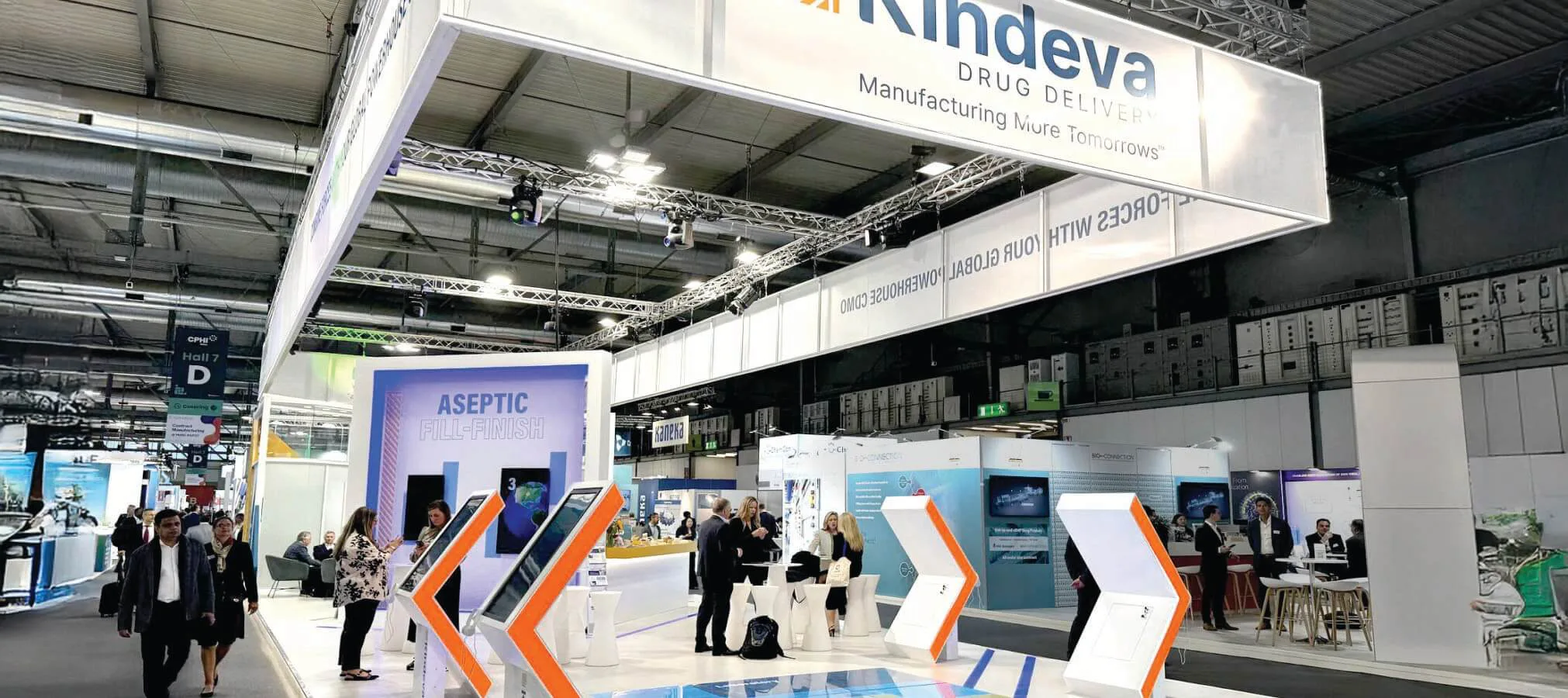 Exhibition booth for Kindeva Drug Delivery at a pharmaceutical trade show. The booth features a modern design with interactive touchscreen displays on white and orange stands. A large banner highlights 'Aseptic Fill-Finish.' Several attendees are engaged in discussions or exploring the booth. The ceiling has an industrial design with exposed ducts and lighting. Other exhibitor booths, including Kindeva, are visible in the background.