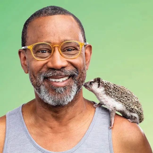 A smiling man with a neatly trimmed beard and glasses wearing a sleeveless gray top, with a small hedgehog perched on his shoulder. The hedgehog appears to be sniffing or interacting with the man's face. The background is a smooth gradient of green.