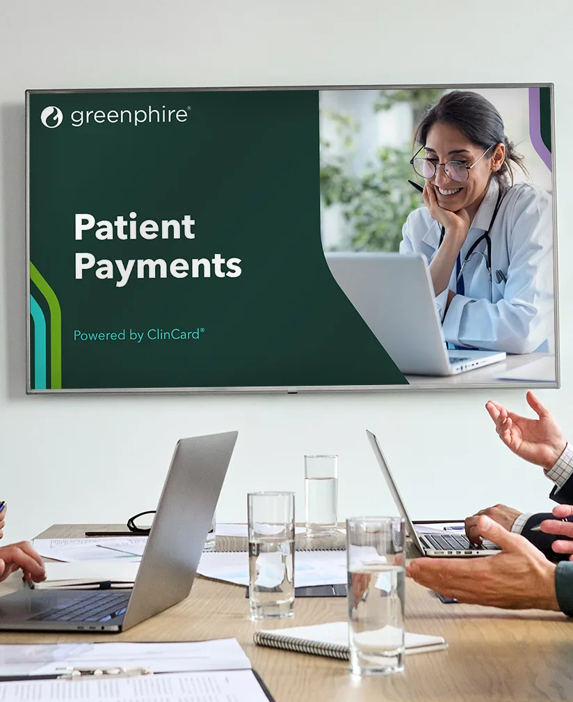 A Greenphire PowerPoint presentation slide displayed on a large TV screen in a conference room. The slide has a dark green background with a white title "Dark Presentation Title" and a subtitle below. An image of a woman, likely a healthcare professional, smiling and working on a laptop is shown on the right side of the slide. The slide uses Greenphire's branding colors and design elements, including a green and purple border. Several people in the meeting are engaged in discussion, seated at a table with laptops, papers, and glasses of water.