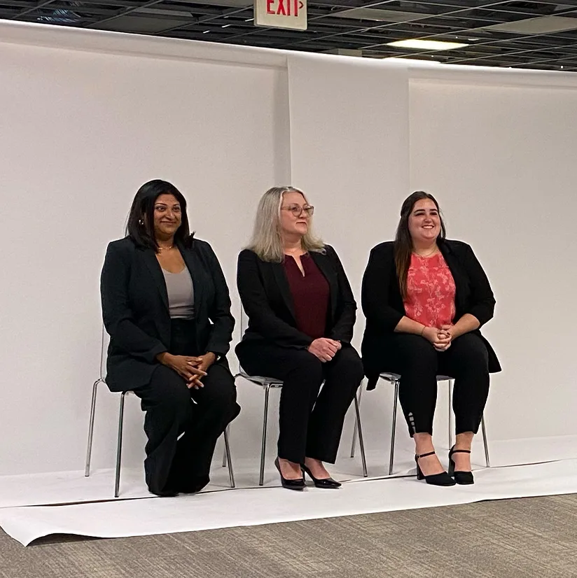 Behind-the-scenes moment from Sterling IRB's 'Big Idea' photoshoot. Three professionally dressed women from the Sterling IRB team are seated against a white backdrop, smiling and posing for the camera. The studio setup, including the backdrop and lighting, is visible, capturing the preparation and teamwork behind the final campaign imagery.