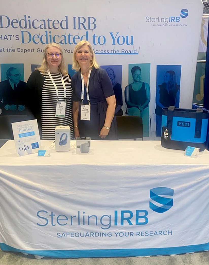 Two women standing behind a Sterling IRB booth at an event or tradeshow. The booth display features the tagline 'Dedicated IRB that's Dedicated to You' and the Sterling IRB logo with the slogan 'Safeguarding Your Research.' Promotional items including a YETI cooler, headphones, and informational brochures are arranged on the table.
