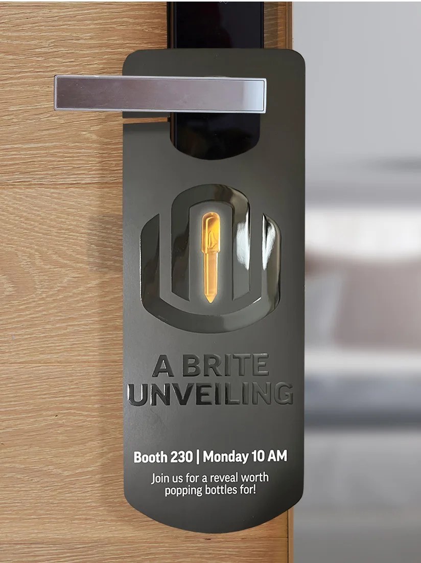 Black door hanger with a glossy embossed Univo IRB logo and a glowing Lite Brite peg in the center, promoting an event called 'A Brite Unveiling.' Text reads: 'Booth 230 | Monday 10 AM. Join us for a reveal worth popping bottles for!' The hanger is displayed on a silver door handle against a wooden door background.
