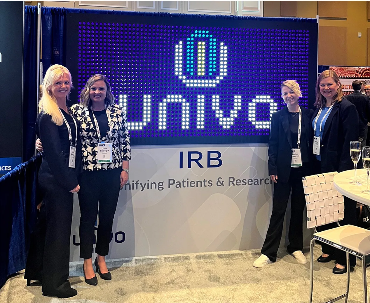Four women from the Univo IRB team stand in front of a Lite-Brite-style display revealing the new Univo IRB name and logo at a trade show. The backdrop features illuminated pegs spelling out 'Univo' with the company shield icon above. The booth display reads 'IRB – Unifying Patients & Research.' Attendees are smiling and dressed in professional attire.