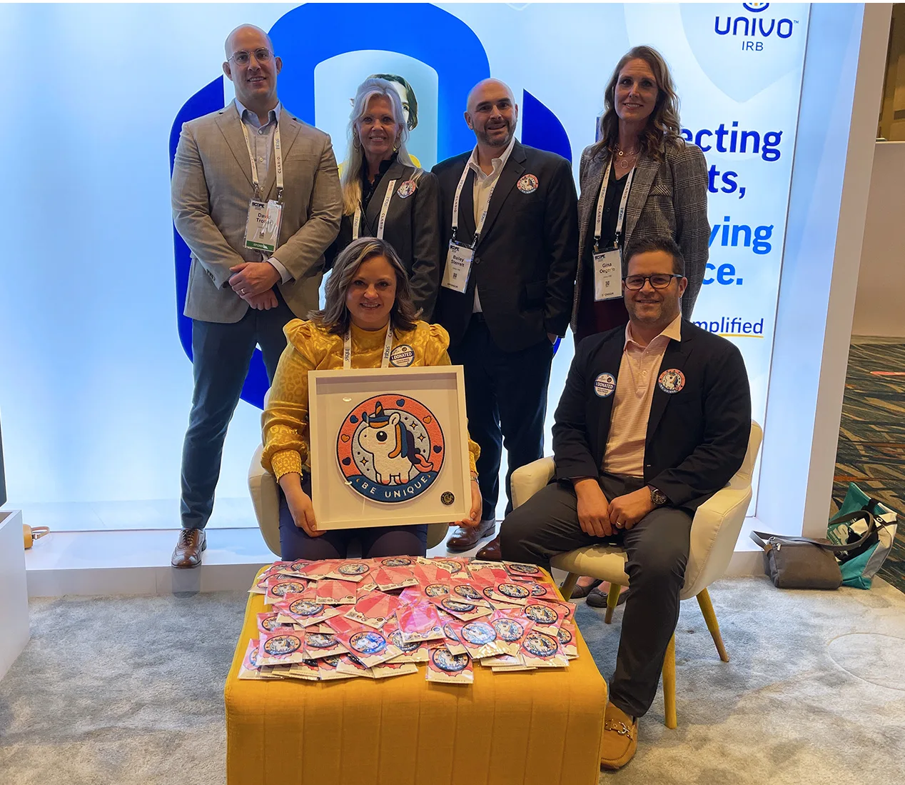 Group photo of the Univo IRB team at a trade show booth, supporting the Oliver Patch Project. Six team members pose in front of a lit backdrop with the Univo logo and slogan 'Protecting Patients, Not Trying Patience.' Two team members are seated, with one holding a large framed patch featuring a unicorn and the words 'Be Unique.' A table in front displays dozens of smaller patches and pink encouragement cards as part of the donation initiative for children undergoing cancer treatment.