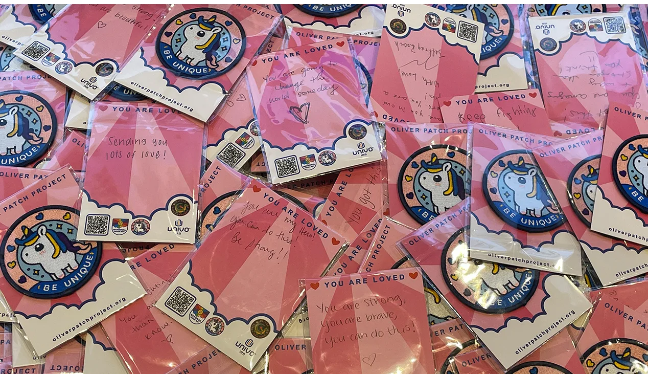 Close-up of dozens of Oliver Patch Project kits arranged on a surface. Each kit includes a colorful unicorn patch with the words 'Be Unique' and a pink card labeled 'You Are Loved,' featuring handwritten encouraging messages like 'You are strong, you are brave,' and 'Sending you lots of love.' The kits also include Univo IRB and Oliver Patch Project logos, and QR codes for more information.