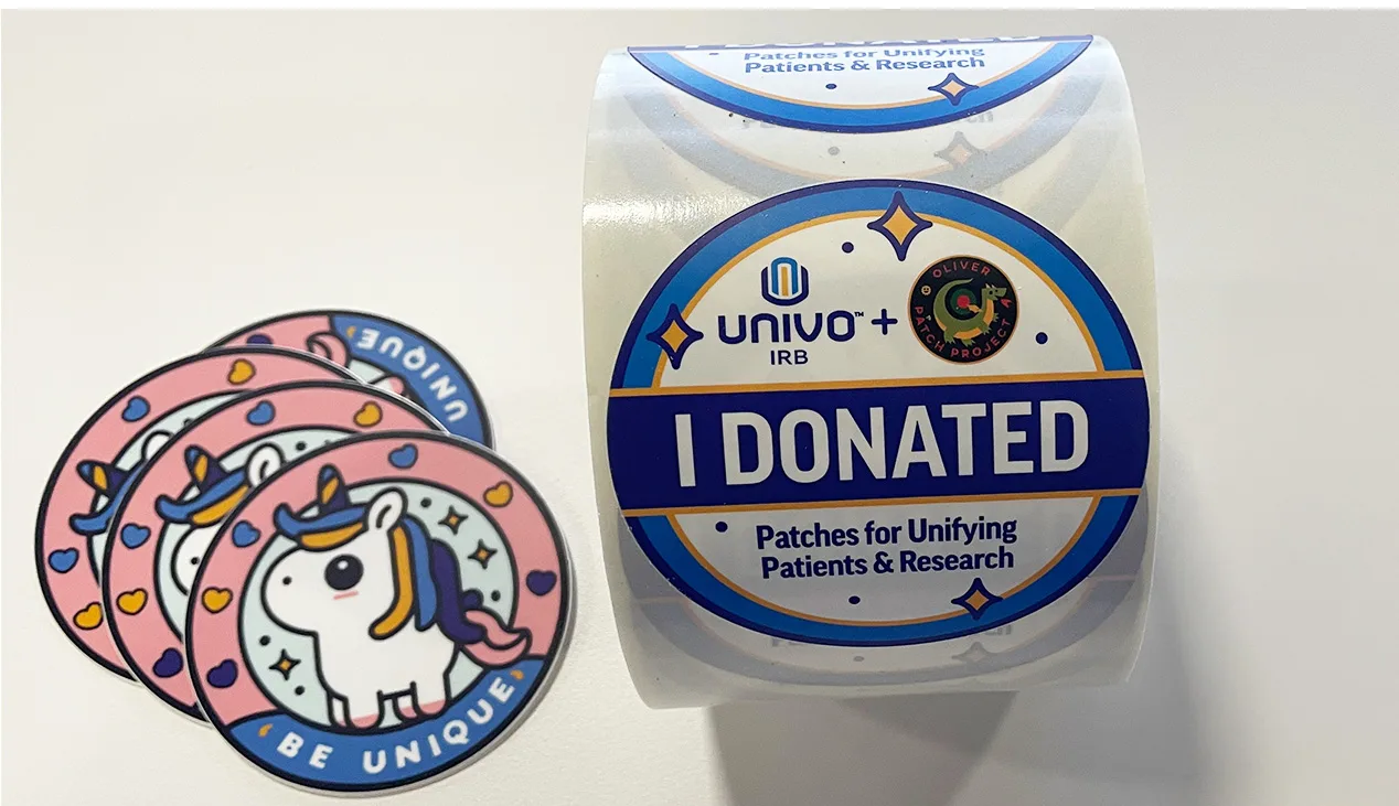 Roll of round 'I Donated' stickers from Univo IRB in partnership with the Oliver Patch Project, labeled 'Patches for Unifying Patients & Research.' Next to the roll are several colorful unicorn patches with the phrase 'Be Unique,' part of a campaign supporting children undergoing cancer treatment.