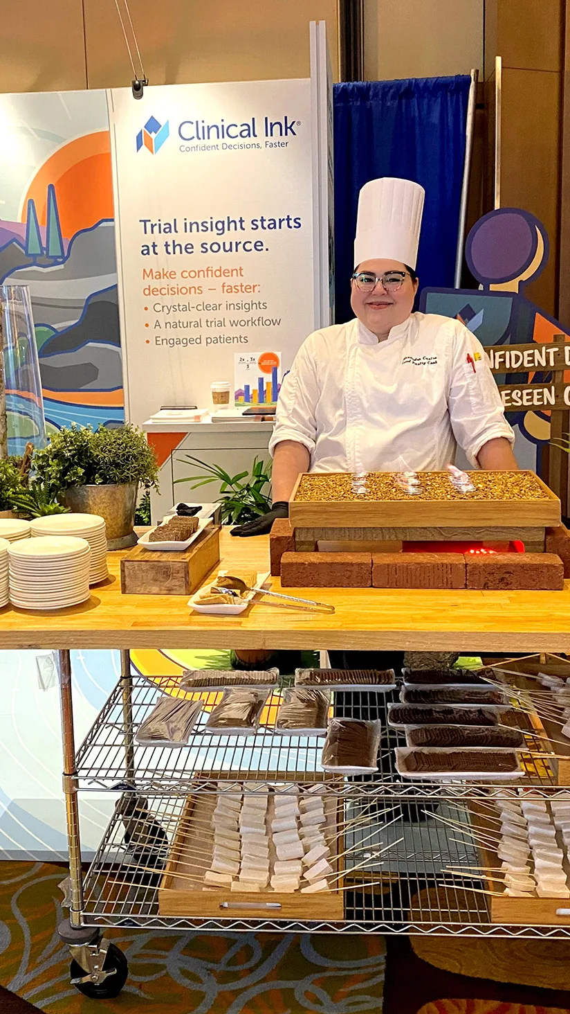 A professional chef in a white uniform and tall toque stands behind a wooden serving station at a Clinical Ink tradeshow booth. Behind her is a branded banner reading “Clinical Ink – Confident Decisions, Faster” and “Trial insight starts at the source. Make confident decisions—faster: • Crystal-clear insights • A natural trial workflow • Engaged patients.” On the table are stacks of small white plates, potted greenery, and a heated wooden tray with bite-sized samples; beneath, a metal cart holds rows of skewered marshmellows and platters ready for guests.