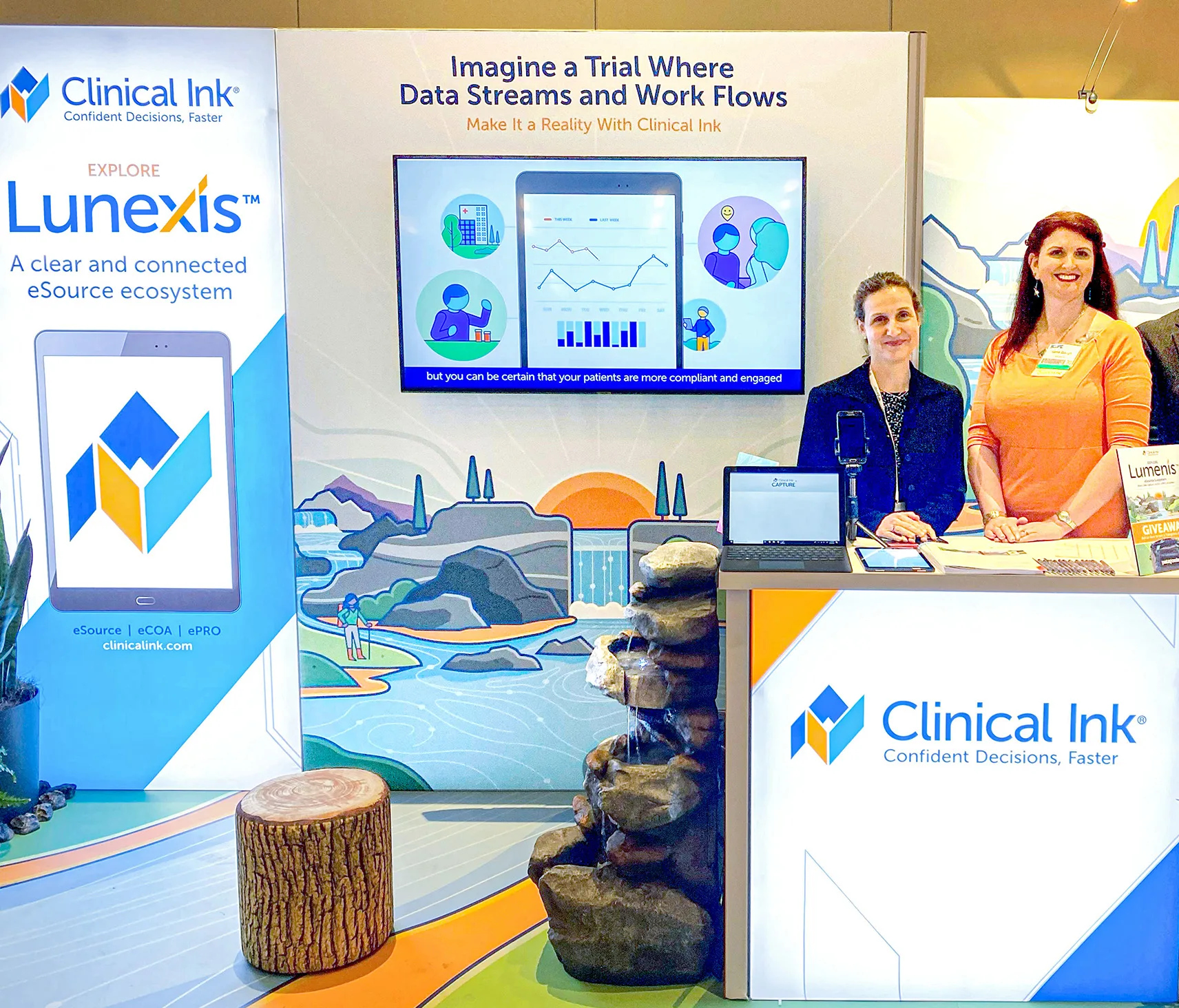 A brightly lit tradeshow booth for Clinical Ink featuring a wraparound backdrop and branded counter. On the left, a tall banner reads “Explore Lunexis™ – A clear and connected eSource ecosystem” above a stylized tablet icon. Centered, a large wall graphic of a river valley and waterfall is topped by the headline “Imagine a Trial Where Data Streams and Work Flows – Make It a Reality With Clinical Ink” and a video screen showing charts and patient icons. In front, a stacked-stone water feature spills into a shallow basin beside a faux-wood stump seat. Two smiling booth staff—a woman in a navy blazer and another in an orange top—stand behind a counter holding laptops, brochures, and a “Lumenis™ Giveaway” sign, ready to engage visitors.