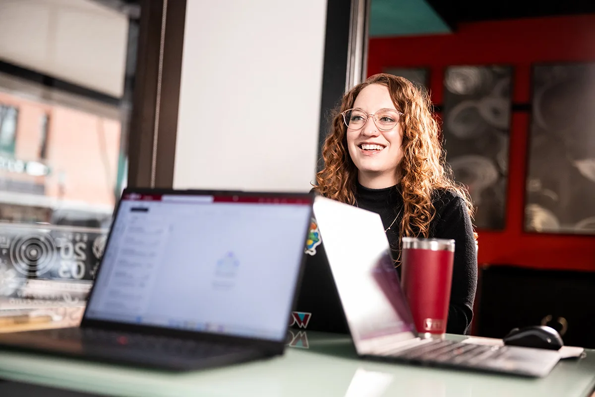 A cozy café-style workspace scene shot over the shoulders of two blurred laptops on a glass-topped table. In the background sits a smiling young woman with curly auburn hair and glasses, wearing a black sweater. A red insulated tumbler stands next to her laptop, and daylight streams through large windows behind her, with red accent walls and framed artwork visible.