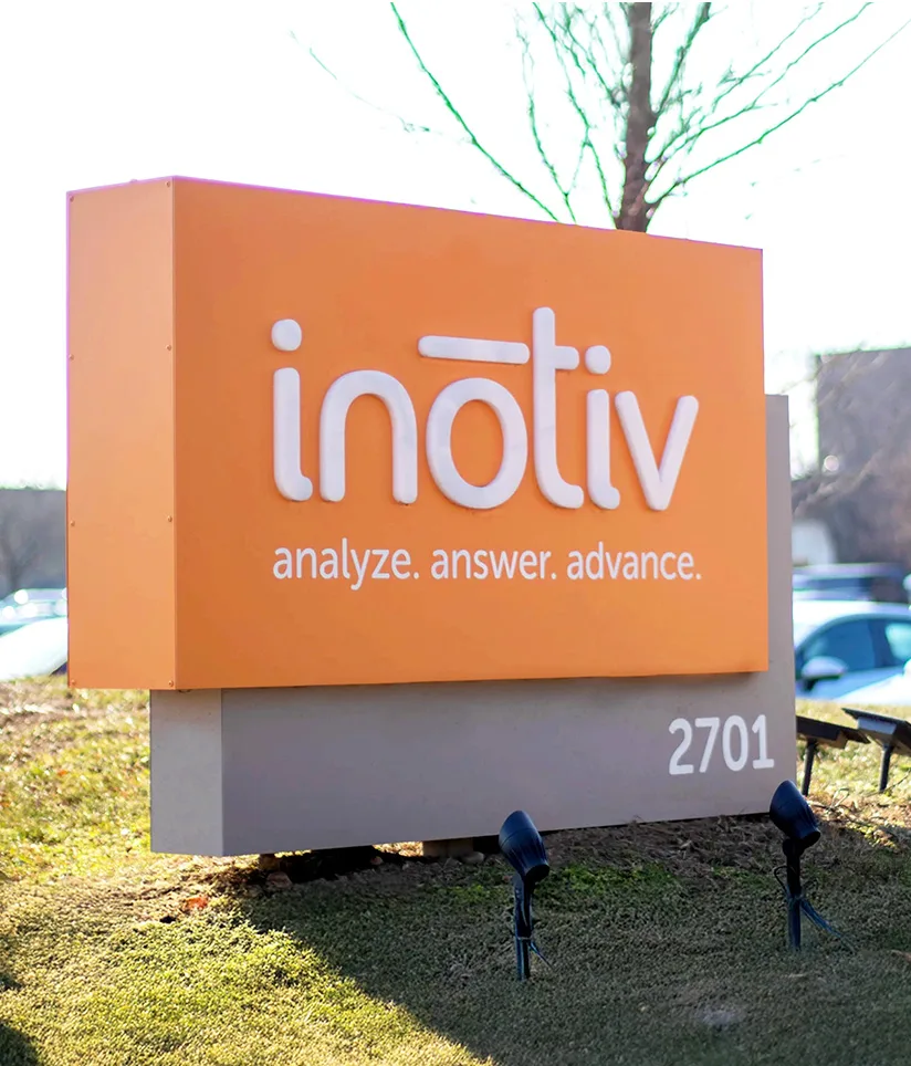 A close-up photograph of a sign featuring the Inotiv logo, displayed in large white letters on an orange background. Below the logo, the slogan "analyze. answer. advance." is written in smaller text. The sign also includes the address "2701" at the bottom