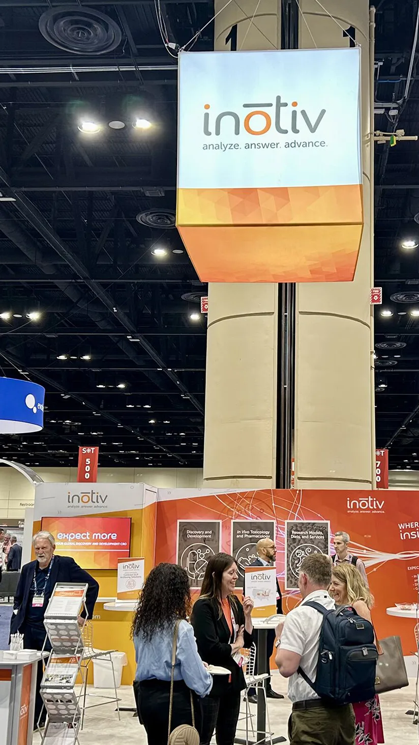 "A tradeshow booth with an overhead sign displaying the 'inotiv' logo and slogan 'analyze. answer. advance.' People are gathered around the booth engaging in conversation, while informational posters and promotional materials are visible. The booth has an orange and white color scheme with the tagline 'Expect More' prominently featured on a screen.