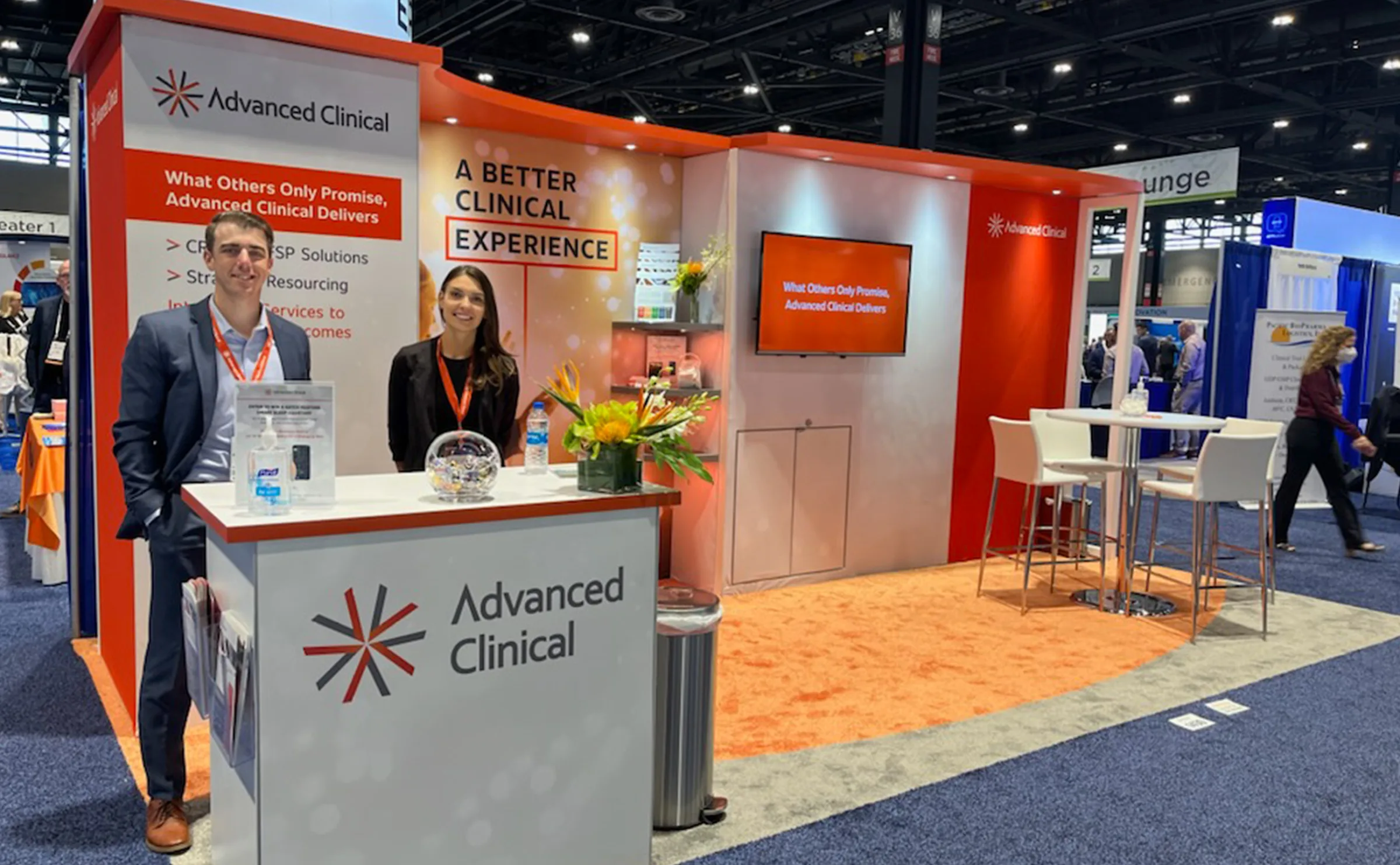 An image showing a trade show booth for Advanced Clinical, with a large orange and white backdrop. The backdrop features their big idea imagery, along with text listing benefits of working with Advanced Clinical. Two professionals are standing behind a counter displaying the Advanced Clinical logo, both smiling.