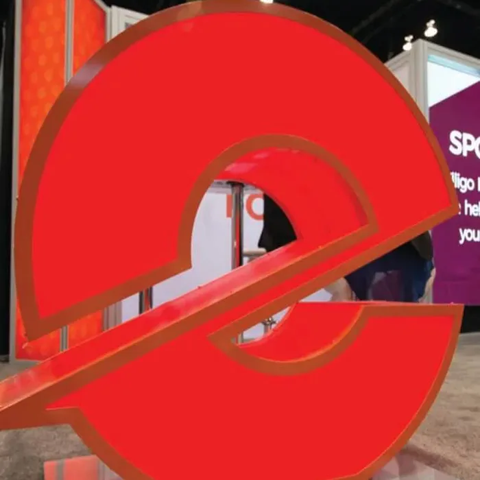 A large, red 3D Elligo logo structure on display at a tradeshow booth, with surrounding signage and exhibit panels in the background.