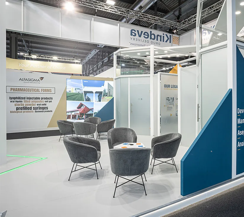 A meeting area within the Kindeva Drug Delivery booth at the CPHI Milan tradeshow, featuring modern gray chairs, small tables with brochures, and glass-walled rooms for private discussions.