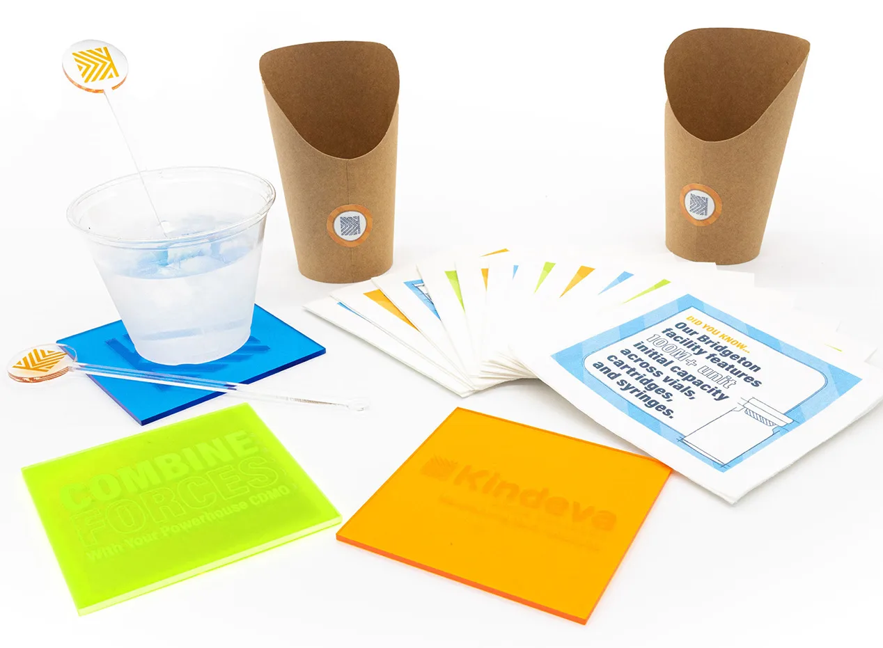 Promotional items from Kindeva's CPHI Milan event, including branded coasters, paper cups, a drink stirrer, and informational napkins featuring facts about their Bridgeton facility.