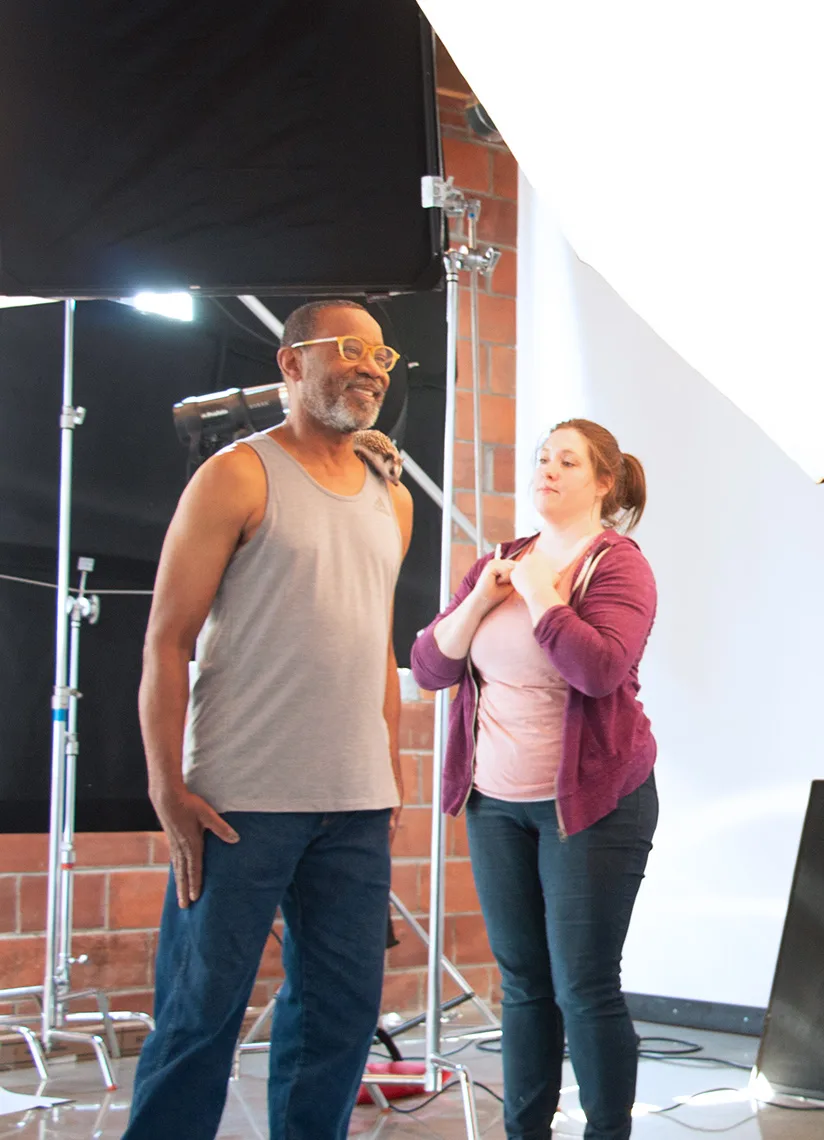 The image shows a behind-the-scenes shot of a photoshoot. A man wearing glasses and a gray tank top stands smiling, with a hedgehog on his shoulder. A woman in a pink hoodie is standing next to him assisting with the shoot setup. The background features lighting equipment and a brick wall, indicating the studio environment.