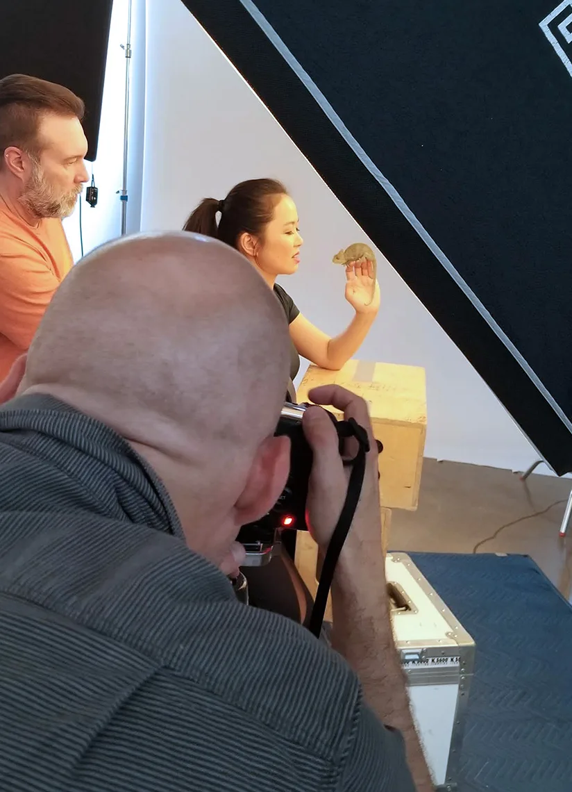The image shows a behind-the-scenes shot of a photoshoot. A woman in a black shirt is holding a chameleon while looking at it. A man in a peach-colored shirt stands next to her assisting. In the foreground, a photographer with a bald head is taking a picture of the scene. The setup includes lighting equipment in the background.