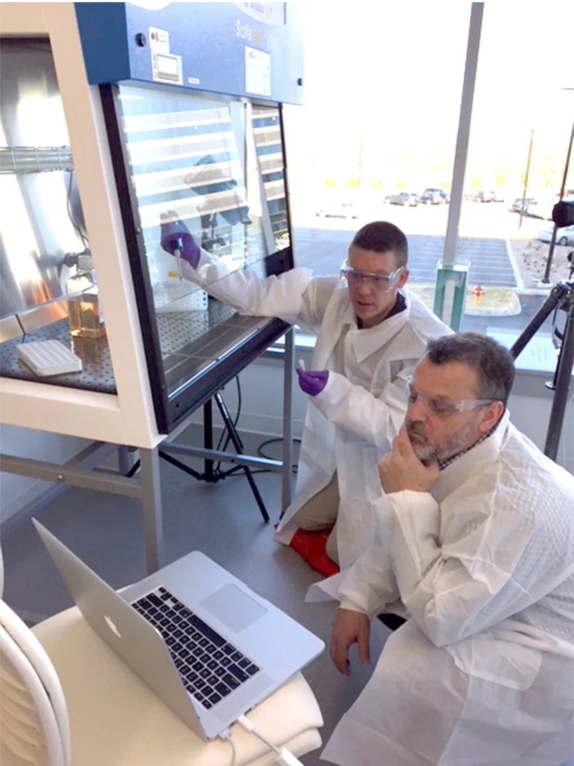 A photoshoot in a laboratory setting with two men in white lab coats and safety glasses. One of them is wearing purple gloves operating equipment inside a biosafety cabinet, while the other is looking at a laptop.