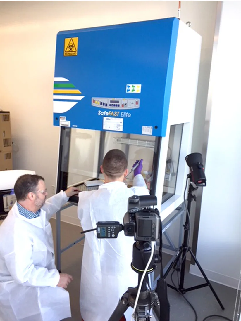 A photoshoot in a laboratory where two men, both wearing white lab coats, are working with equipment inside a biosafety cabinet labeled 'SafeFAST Elite.' One of them is handling materials inside the cabinet, while the other is observing. A camera and tripod are positioned nearby to capture the scene.