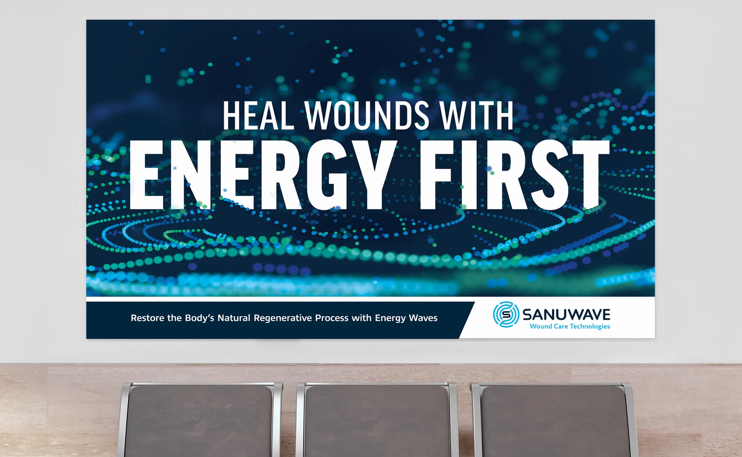An image of a large banner displaying Sanuwave's Big Idea concept. The banner features the slogan 'HEAL WOUNDS WITH ENERGY FIRST' in bold, white letters against a backdrop of abstract blue and green dots. The bottom of the banner includes the tagline 'Restore the Body’s Natural Regenerative Process with Energy Waves' and the Sanuwave logo. This visual emphasizes Sanuwave's focus on using energy-based technologies for wound care.