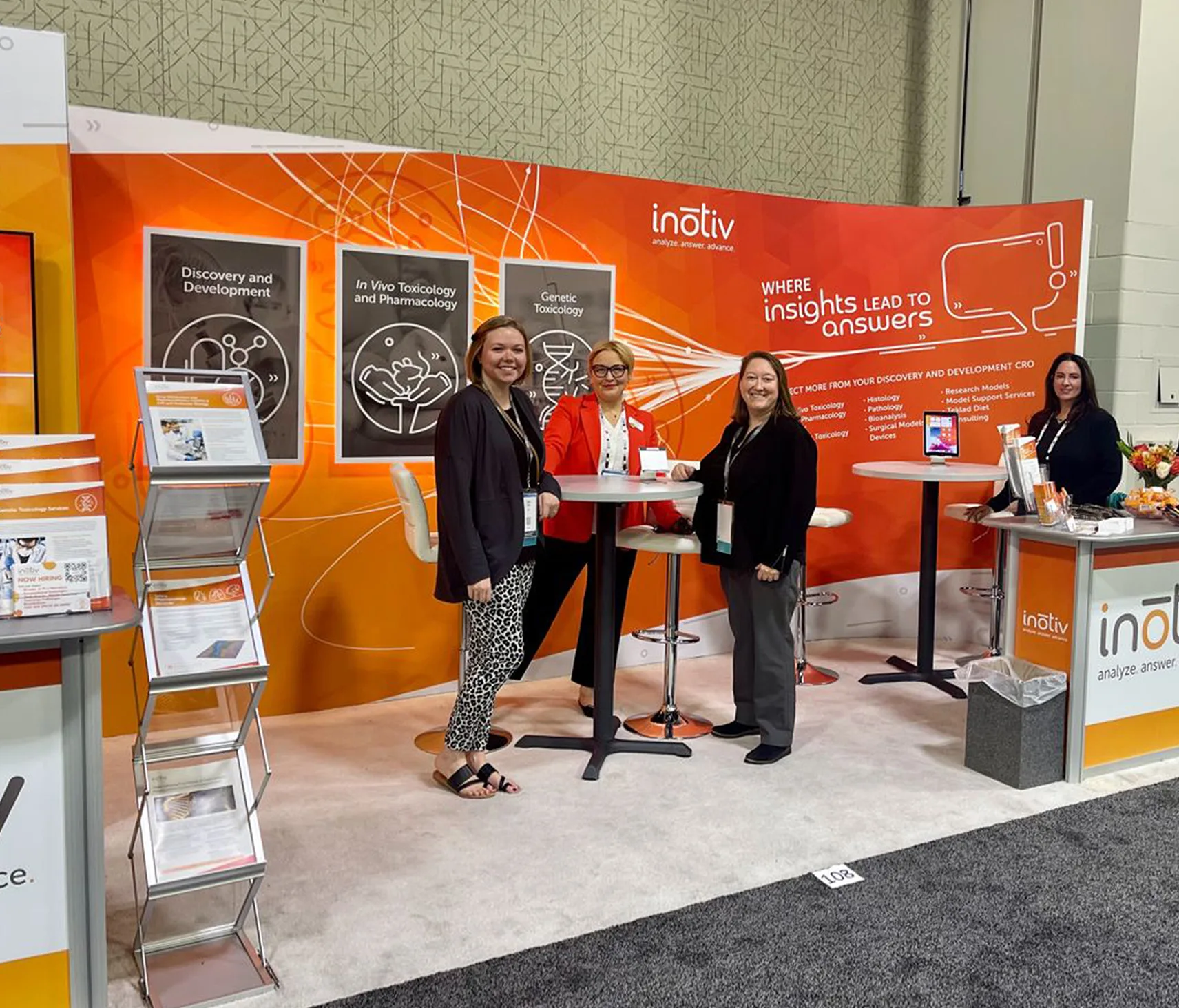 "Four women stand together at an Inotiv tradeshow booth, smiling and interacting. The booth features an orange and white color scheme with the Inotiv logo and tagline 'Where Insights Lead to Answers.' Behind them are three posters showcasing services related to Discovery and Development, In Vivo Toxicology and Pharmacology, and Genetic Toxicology. A display stand with informational brochures is also visible.