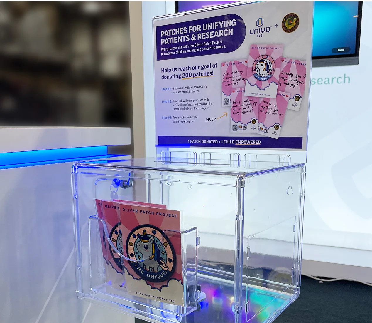 Clear donation box at a Univo IRB booth for the 'Patches for Unifying Patients & Research' initiative in partnership with the Oliver Patch Project. The sign encourages attendees to write encouraging notes that will be sent to children undergoing cancer treatment, with a goal of donating 200 patches. The display includes sample patches with messages like 'You are loved' and 'Wishing you magic, rainbows, and hugs.' A sign explains that one patch donated equals one child empowered.