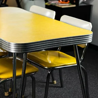 A retro-style dining set featuring a yellow laminate tabletop with rounded corners and chrome edging, paired with matching yellow metal chairs that have tubular steel legs, all set on a dark carpeted floor.