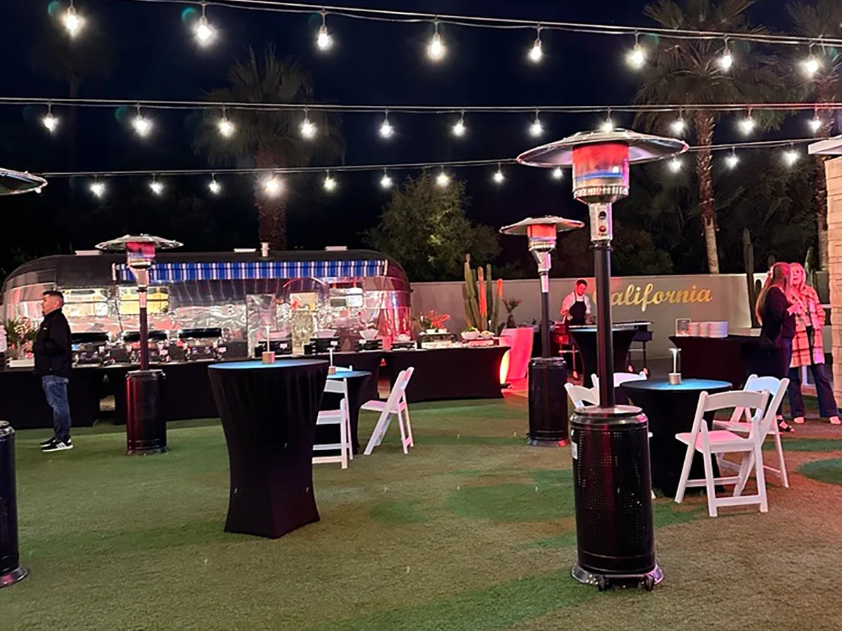 Outdoor evening event setup with string lights overhead, patio heaters, and a buffet area in front of a retro-style food truck. A few attendees stand and chat around high-top and round tables on a grass surface. The word 'California' is painted on the wall in the background.