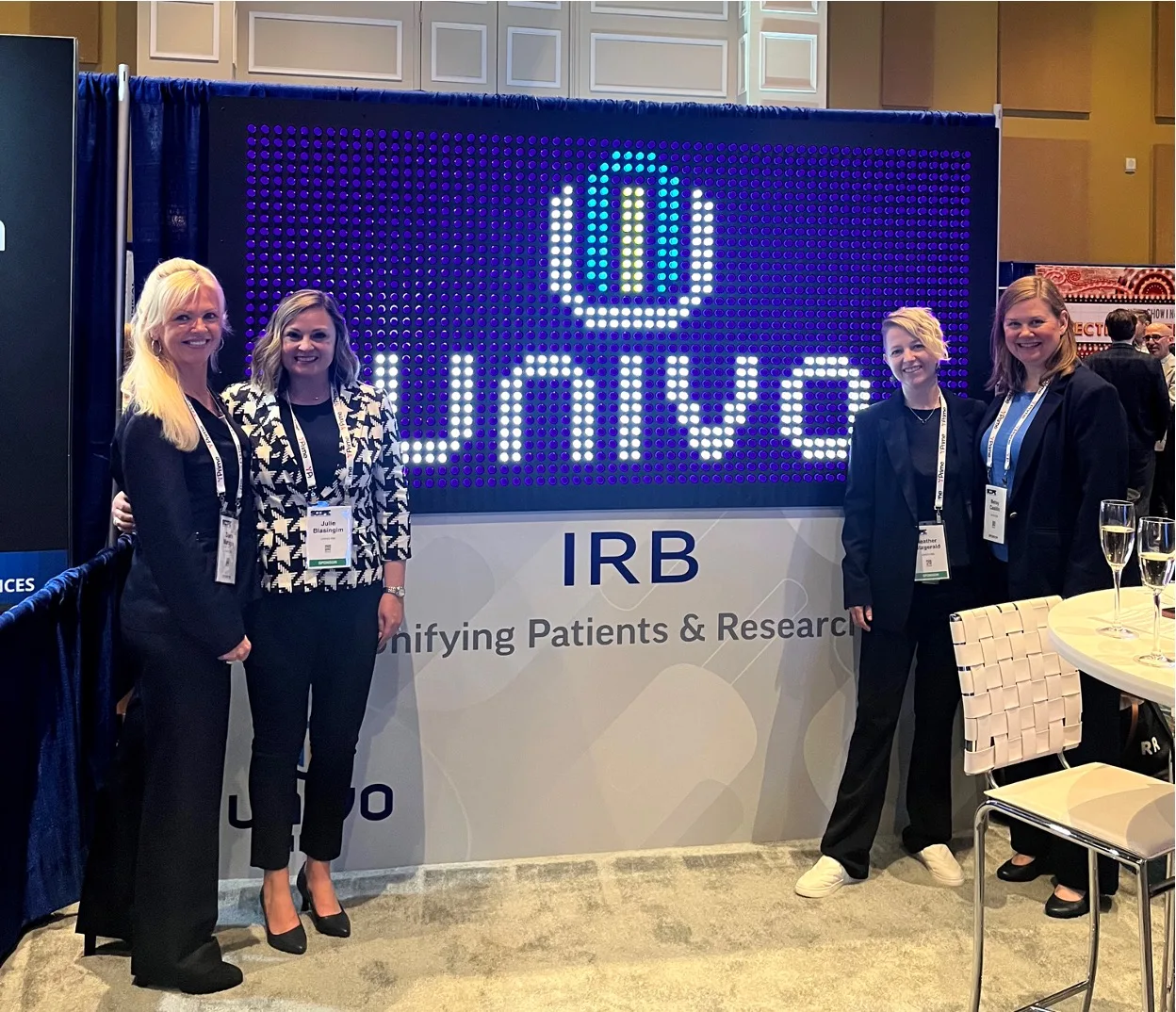 Four women standing and smiling in front of a Univo IRB display booth at a conference. The backdrop features a bright, illuminated Univo logo with the tagline 'Unifying Patients & Research.' All four women are wearing business attire and event badges.