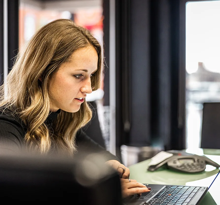 A young woman with wavy blonde hair intently working on a laptop in a modern office setting, with a conference phone and another laptop visible on the table.