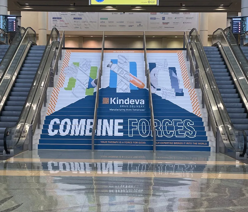 A large staircase graphic at the AAPS tradeshow used as a guerrilla marketing tactic for Kindeva’s brand launch, featuring the message “Combine Forces” and branded visuals across multiple steps between escalators.