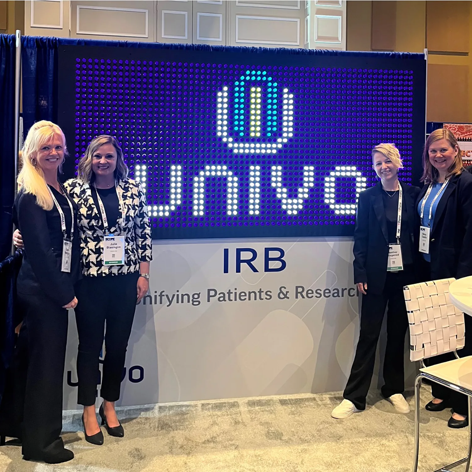 Four team members standing in front of the Univo IRB booth featuring a bright LED-style logo wall and tagline about unifying patients and research, reflecting life science marketing at a conference.