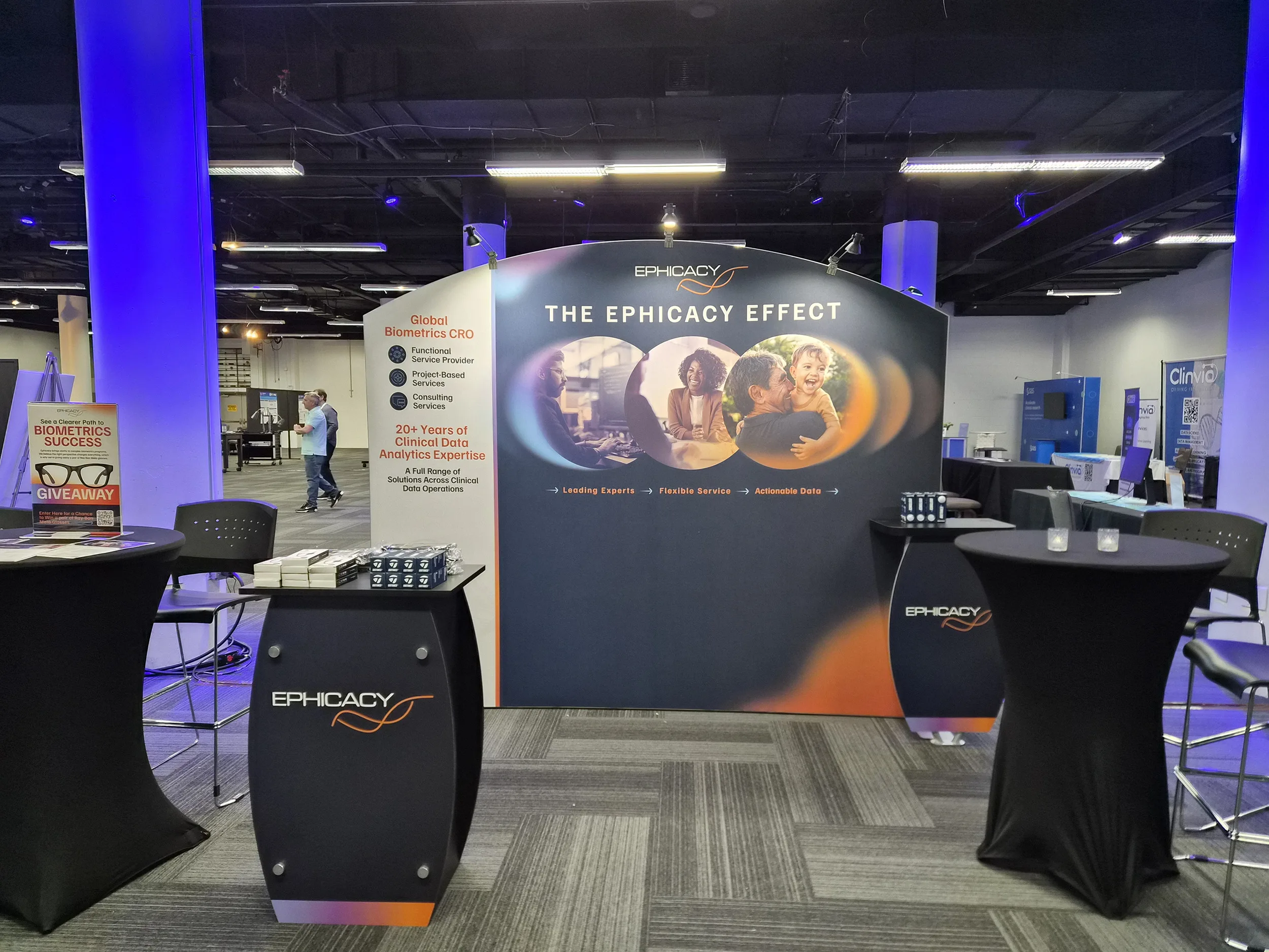 Trade show booth for Ephicacy with a large branded backdrop reading The Ephicacy Effect, a front table displaying brochures and promotional items, and surrounding conference attendees and booths