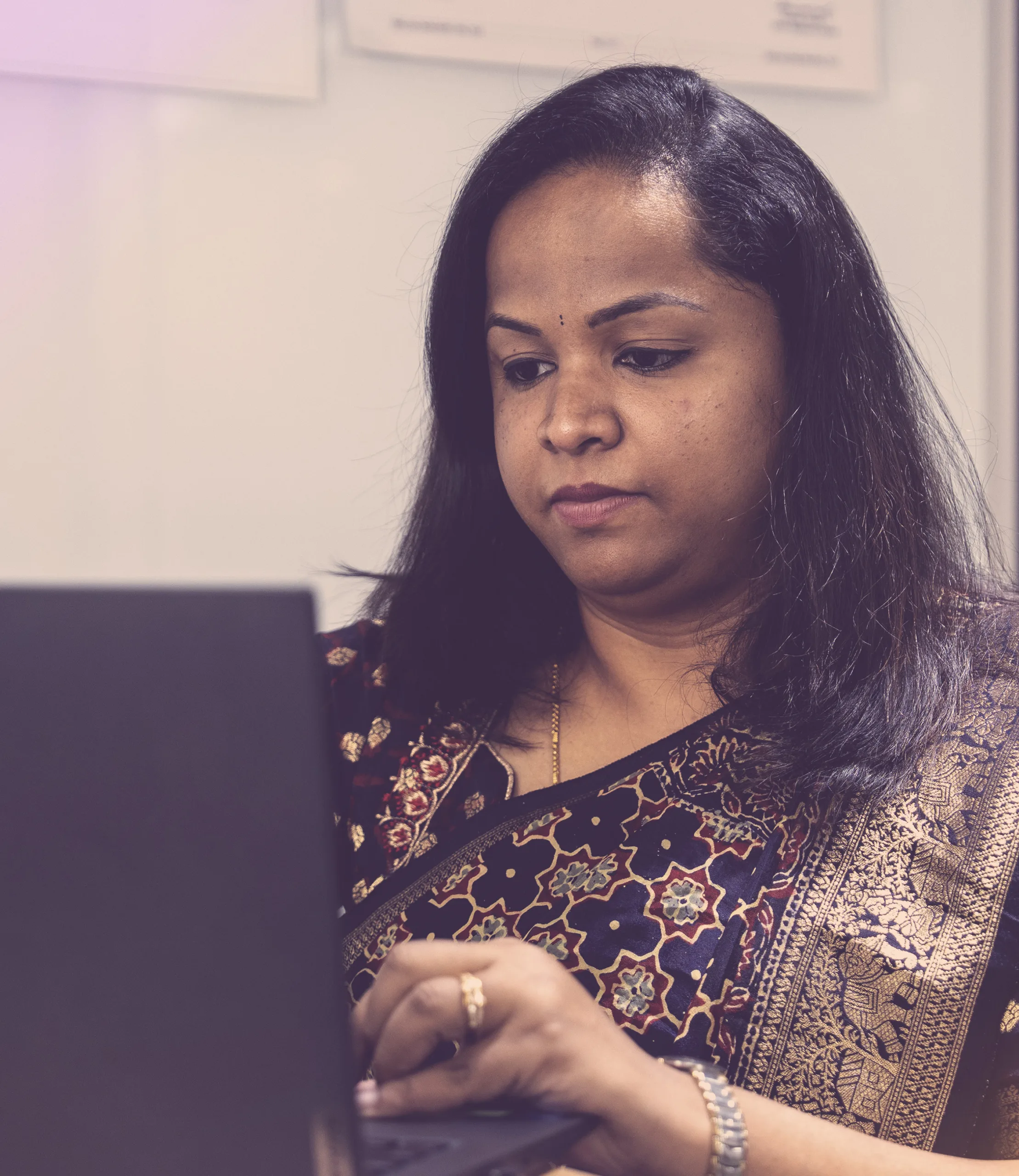 Woman wearing patterned attire working on a laptop with a focused expression in an office setting