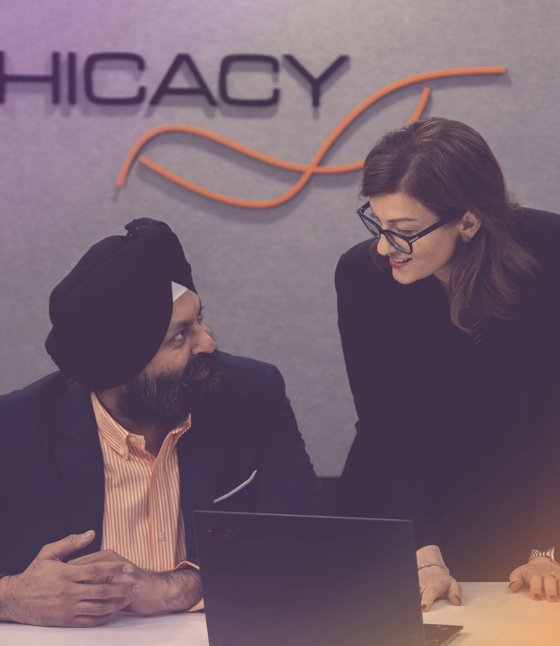Two professionals collaborating at a desk with a laptop, one seated wearing a turban and the other standing and smiling, with Ephicacy branding visible on the wall behind them