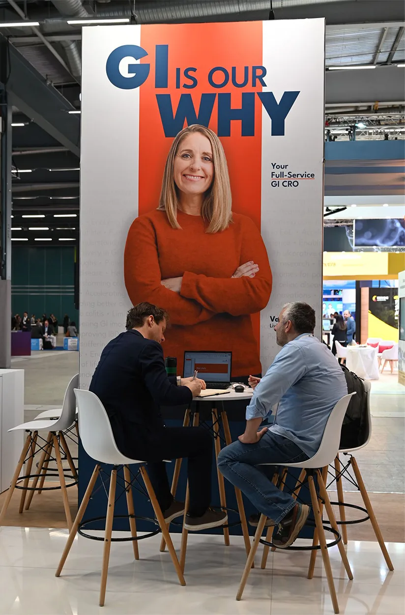 Large vertical booth panel with woman in orange sweater and text GI is our why above two men seated at table in discussion